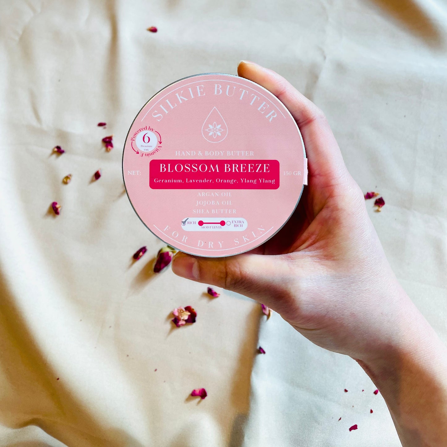 Hand holding a pink container labeled 'Silkie Butter Blossom Breeze' on a light fabric background with scattered dried flowers.